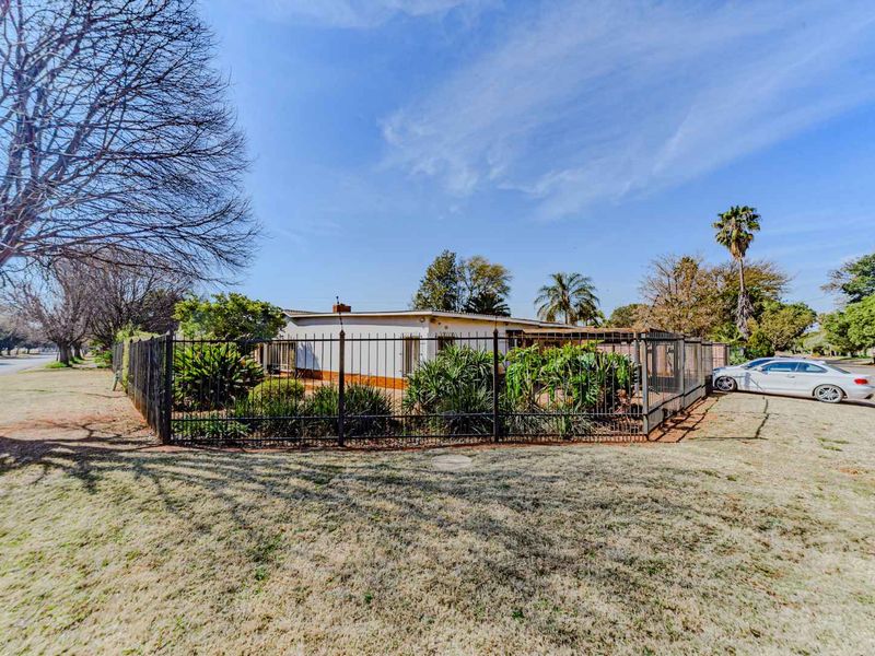 A corner-lot residential property featuring a single-story home enclosed by metal perimeter fencing with a paved driveway and established garden area.