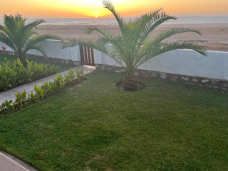 A scenic view of the ocean at sunset, featuring a well-maintained garden, stone boundary wall, and direct access to the beach area.