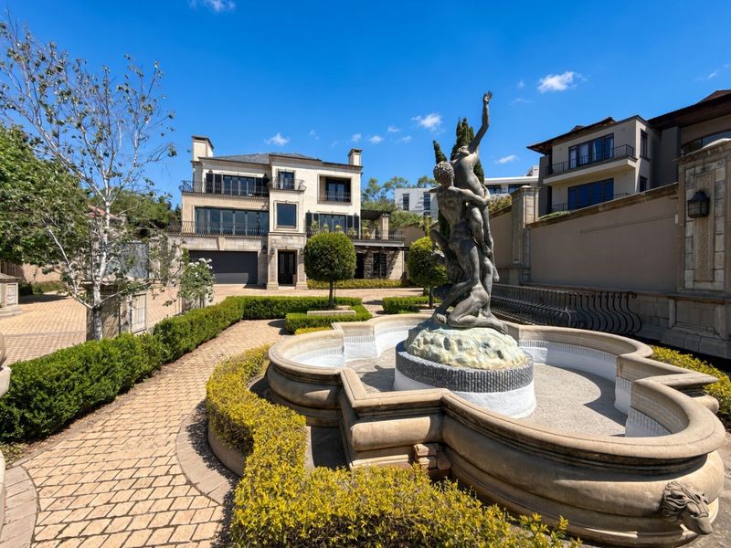 A beautifully landscaped rear garden featuring a central stone fountain with a sculpture, paved pathways, and a multi-story luxury residence in the background.