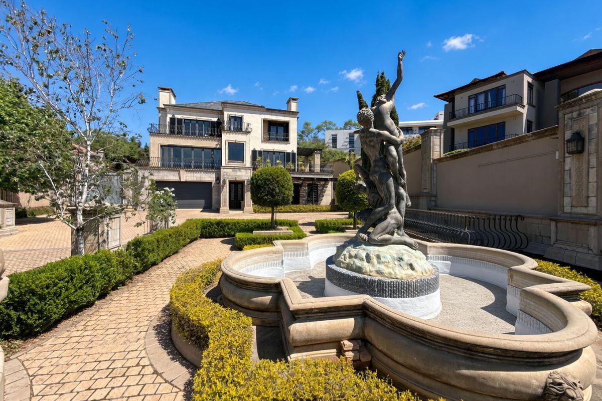 A beautifully landscaped rear garden featuring a central stone fountain with a sculpture, paved pathways, and a multi-story luxury residence in the background.