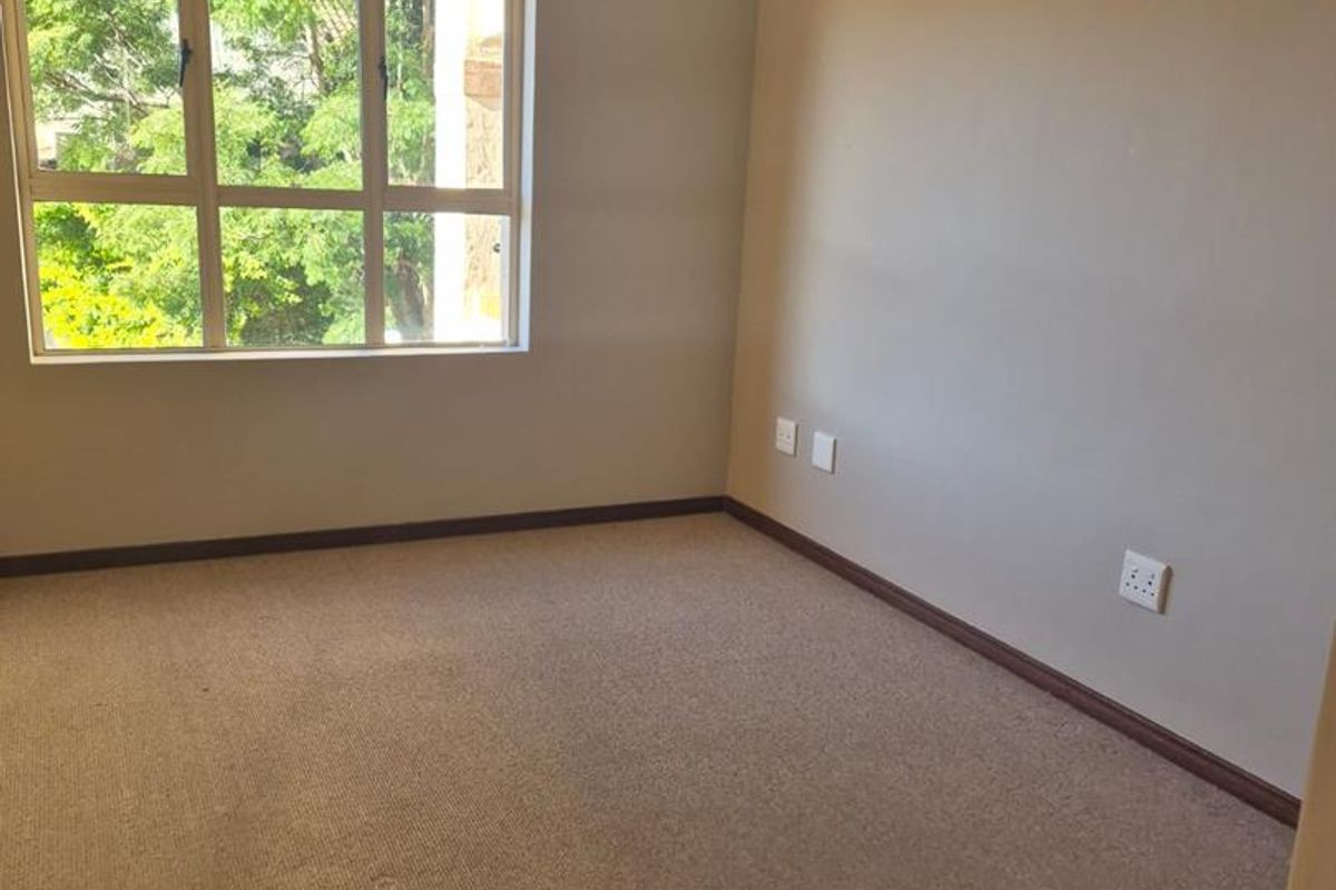 A vacant bedroom featuring neutral wall tones, carpeted flooring, a central ceiling light fixture, and a large window providing natural light.