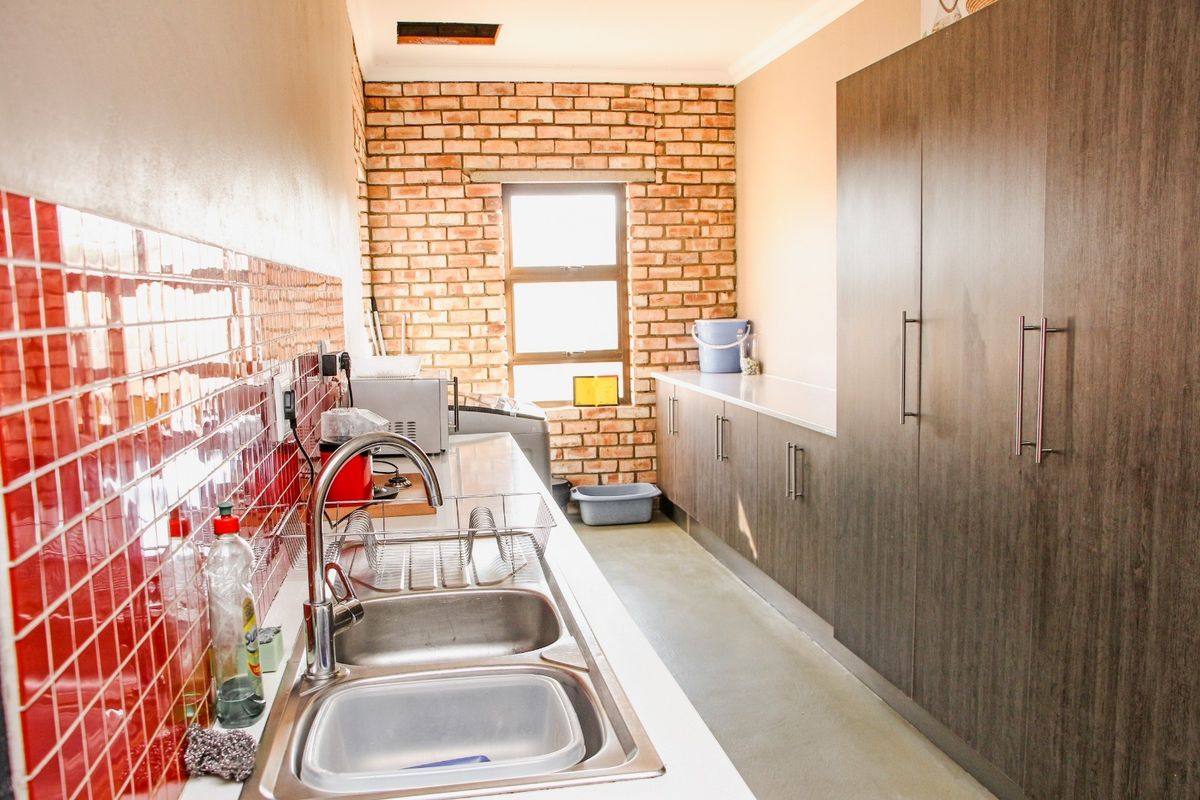 Functional scullery featuring a double stainless steel sink, red tiled splashback, ample built-in cabinetry, and an exposed brick feature wall.