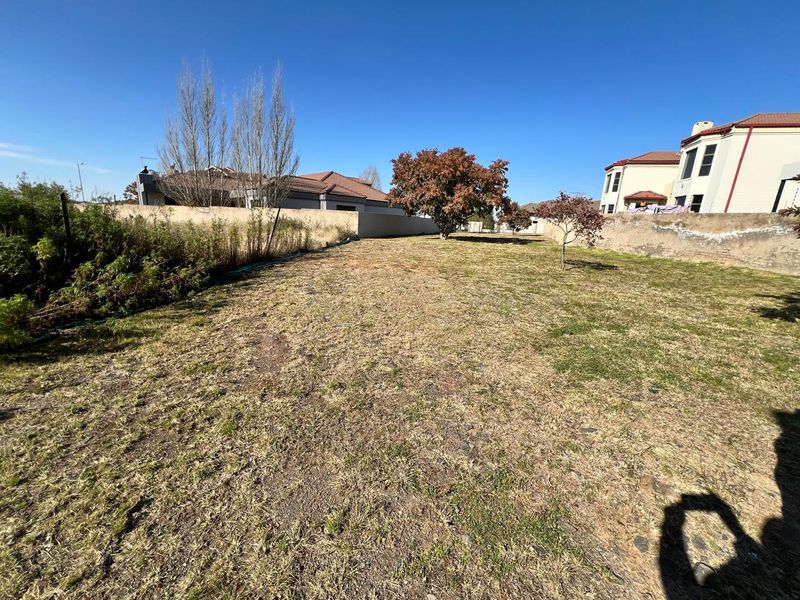 A vacant residential plot featuring a large open grass area enclosed by perimeter boundary walls and mature trees.