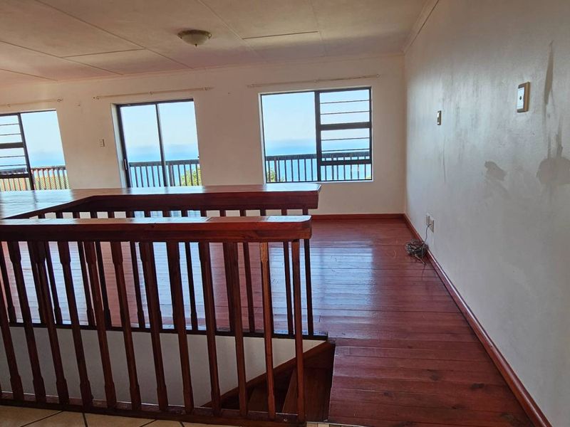 An empty living area featuring timber flooring, a wooden balustrade, and large windows with ocean views, requiring cosmetic wall repairs.
