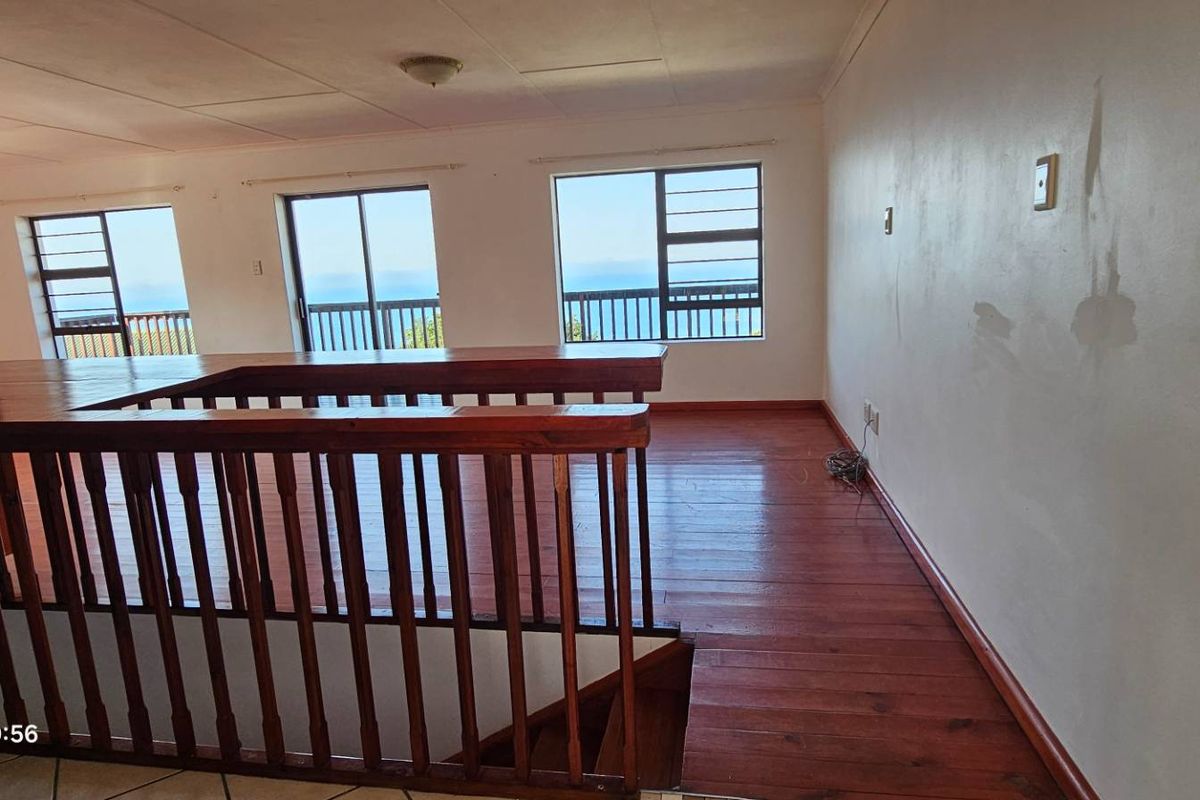An empty living area featuring timber flooring, a wooden balustrade, and large windows with ocean views, requiring cosmetic wall repairs.