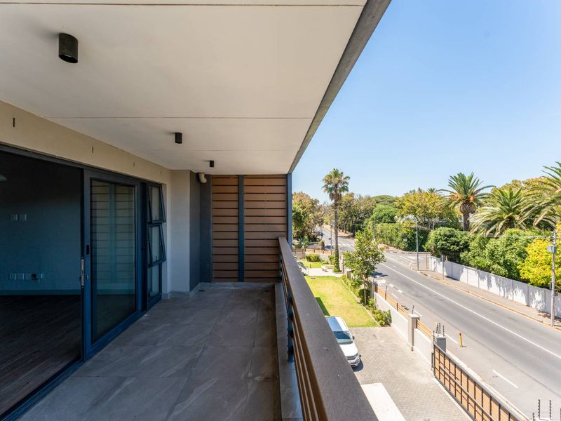 A modern, covered balcony featuring tiled flooring, recessed lighting, and a metal balustrade, offering an elevated view of the street and surrounding greenery.