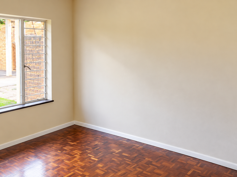 A bright, empty bedroom featuring well-maintained parquet flooring and a window with security bars, offering a clean and neutral space ready for personalization.