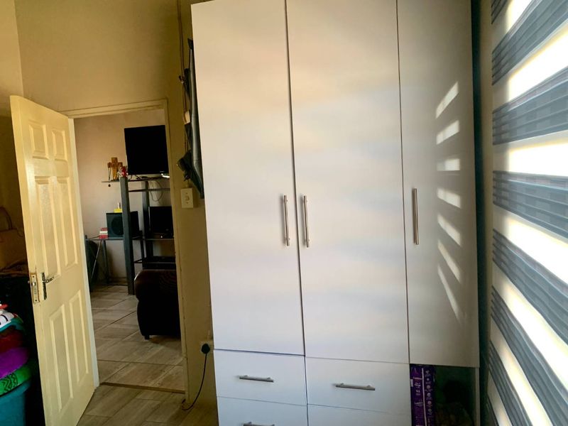 A bedroom featuring a white built-in wardrobe with metal handles, tiled flooring, and modern roller blinds, with a view into an adjacent living area.