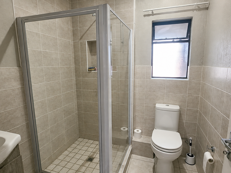 A clean, functional bathroom featuring a glass shower enclosure, tiled walls, a modern toilet, and a wall-mounted basin with natural light from a window.