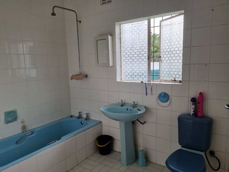 A dated bathroom featuring blue sanitaryware, including a bathtub, pedestal sink, and toilet, with white wall tiling and a window with security bars.