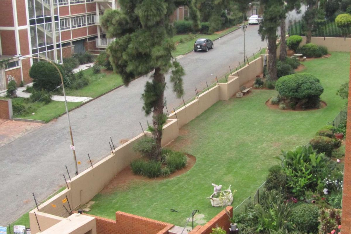 Elevated view overlooking the apartment complex grounds, featuring landscaped lawns, mature trees, and a perimeter wall with security fencing.