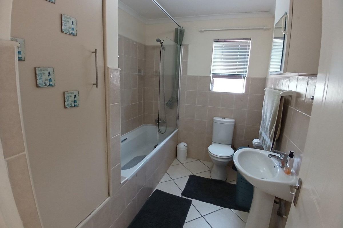 A functional bathroom featuring a bathtub with a glass shower screen, pedestal basin, toilet, and neutral wall and floor tiling.