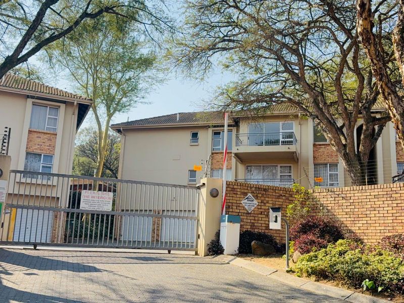 Front view of the apartment complex entrance featuring a secure gated access point, brick boundary walls, and established trees.
