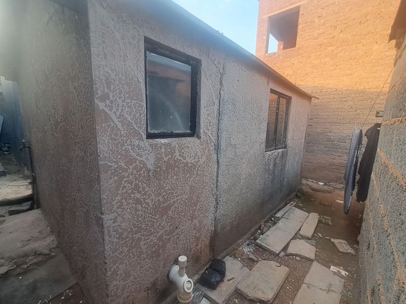 Rear exterior view of a small structure showing textured wall finish, metal window frames, and exposed external plumbing.