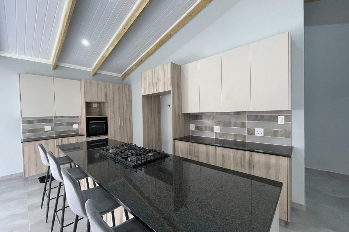 Modern kitchen featuring light wood cabinetry, dark granite countertops, a central island with a gas hob, and exposed timber ceiling beams.