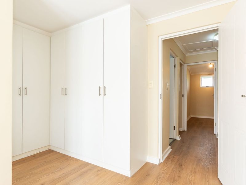 A bright, empty bedroom featuring clean white built-in cupboards, modern laminate flooring, and neutral wall finishes, leading into a connecting hallway.