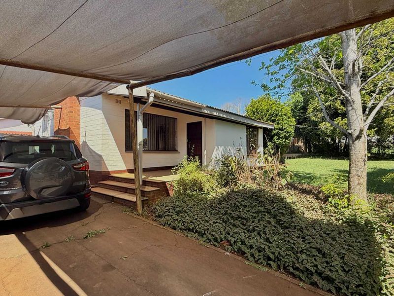 Front view of a single-story brick house featuring a covered carport, paved driveway, and established garden area.