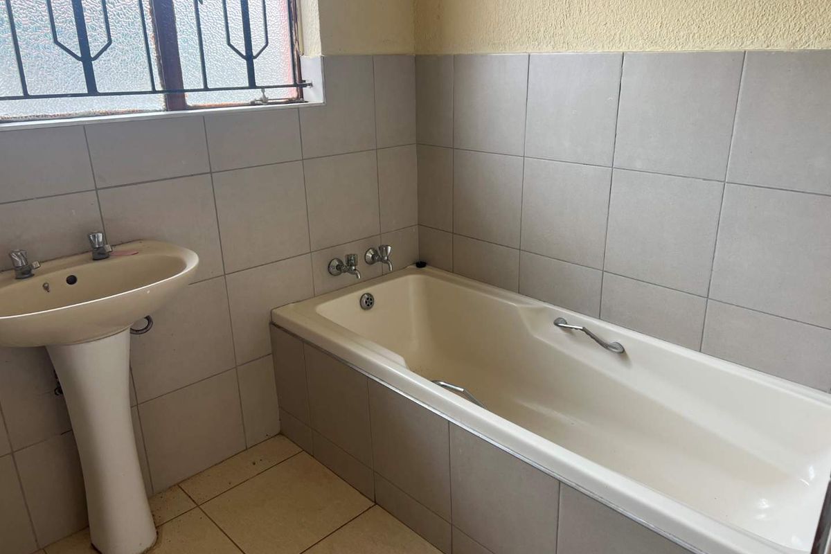 A functional bathroom featuring a pedestal basin, a built-in bathtub with tiled surround, and a security-barred window.