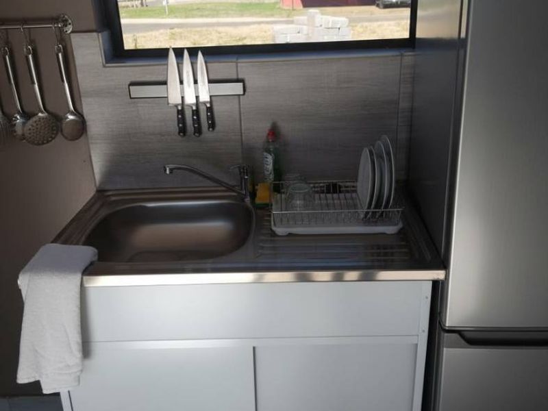 A compact kitchen area featuring a stainless steel sink, tiled splashback, and functional sliding door cabinetry under a horizontal window.