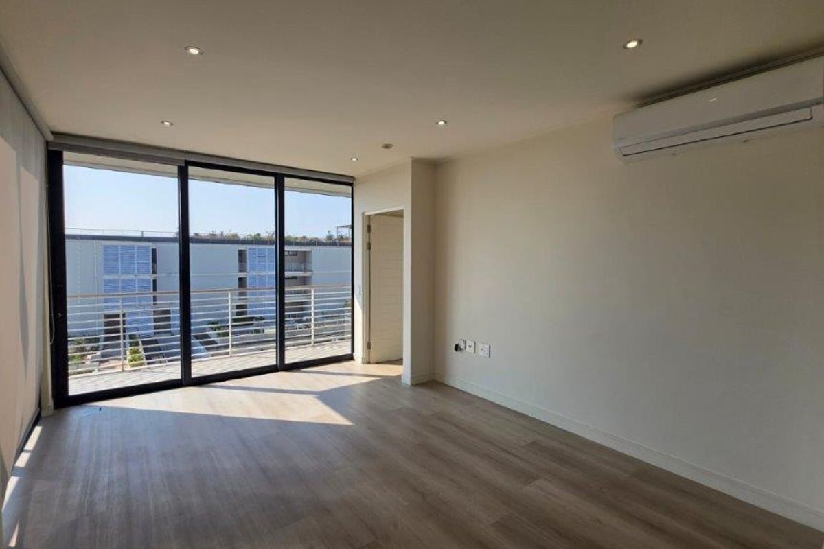 A bright, modern, and empty living room featuring laminate flooring, recessed lighting, a wall-mounted air conditioner, and large sliding doors leading to a balcony.