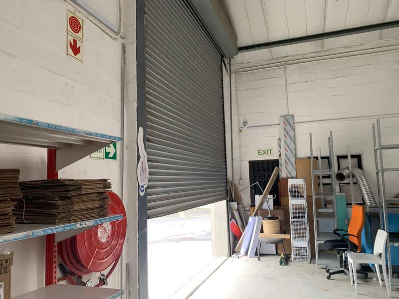 Industrial utility space featuring a large roller shutter door, exposed ceiling structure, and fire safety equipment, suitable for storage or workshop operations.