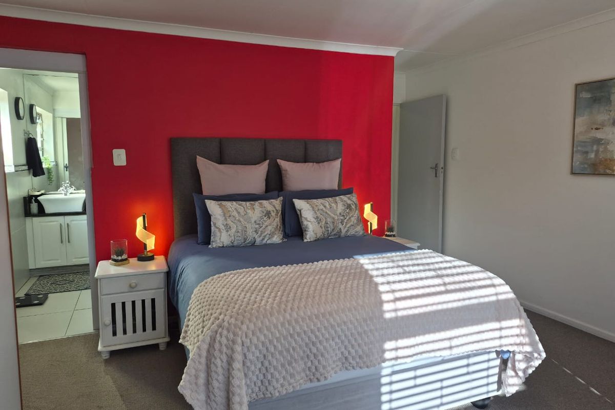 A well-lit bedroom featuring a bold red accent wall, neutral carpeting, and direct access to an adjoining en-suite bathroom.