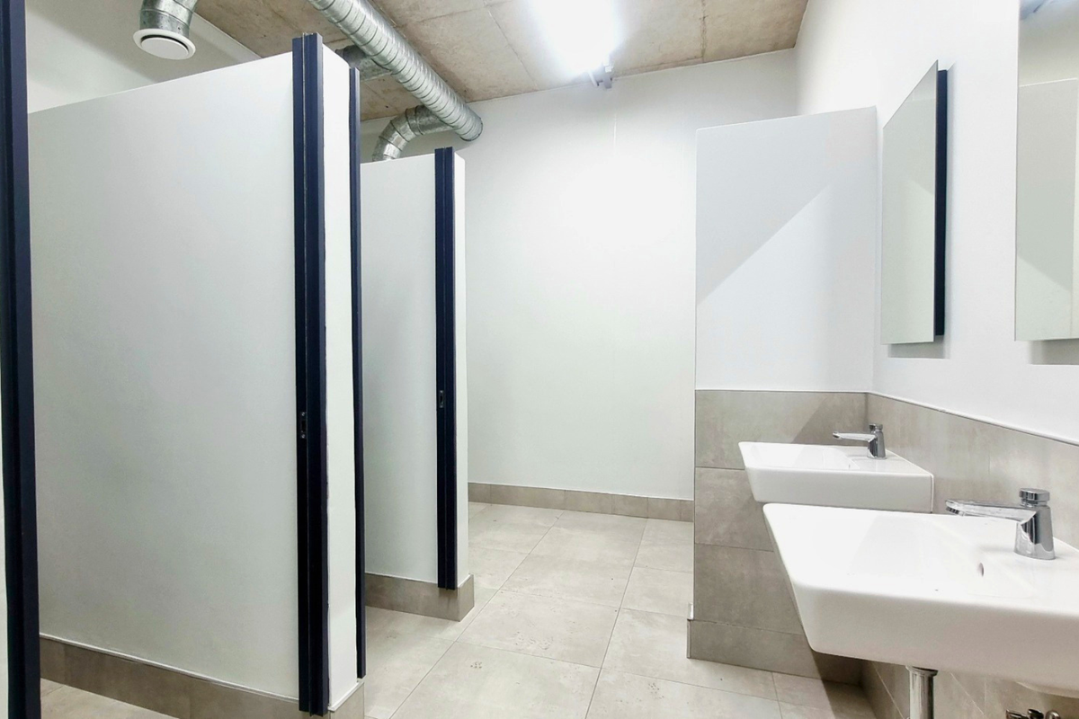 Modern industrial-style communal bathroom featuring partitioned toilet stalls, wall-mounted sinks with chrome fixtures, and exposed ceiling ductwork.
