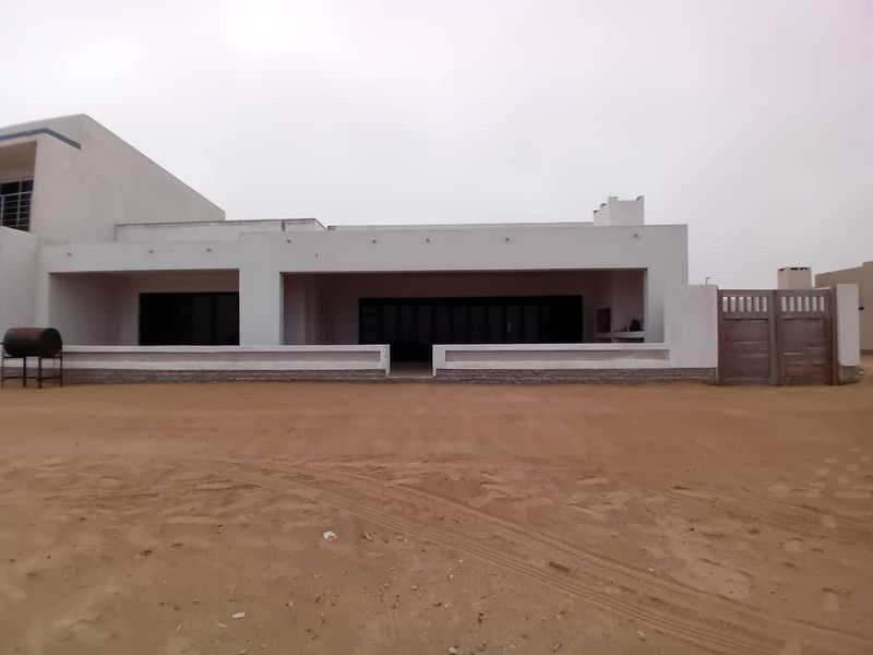 Rear view of a modern house featuring a covered patio, large sliding glass doors, and a built-in braai area, set on an undeveloped sandy plot.