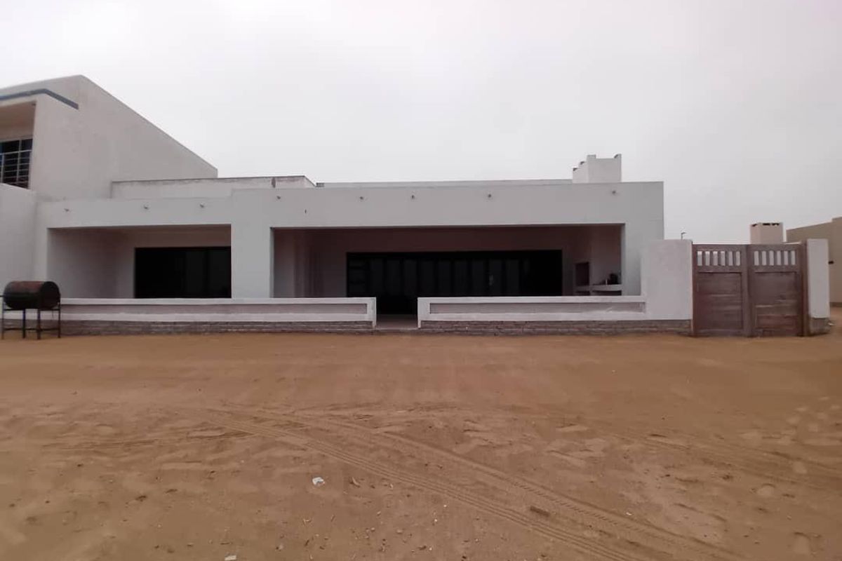 Rear view of a modern house featuring a covered patio, large sliding glass doors, and a built-in braai area, set on an undeveloped sandy plot.