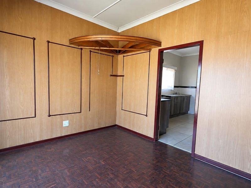 An empty living room featuring wood veneer wall paneling and parquet flooring, with a doorway leading into an adjacent kitchen area.