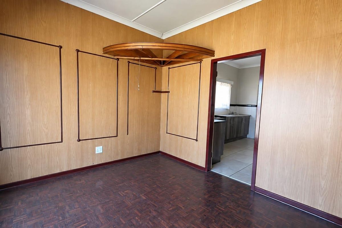 An empty living room featuring wood veneer wall paneling and parquet flooring, with a doorway leading into an adjacent kitchen area.