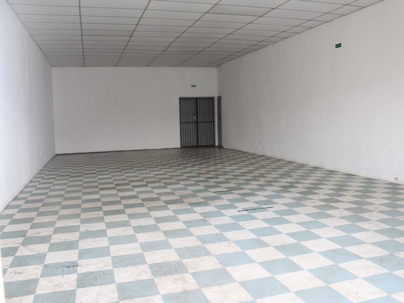 An empty, open-plan retail space featuring a suspended ceiling, checkered vinyl flooring, and a secure metal entrance gate.