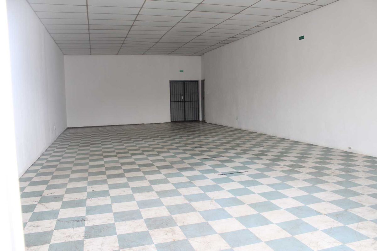 An empty, open-plan retail space featuring a suspended ceiling, checkered vinyl flooring, and a secure metal entrance gate.