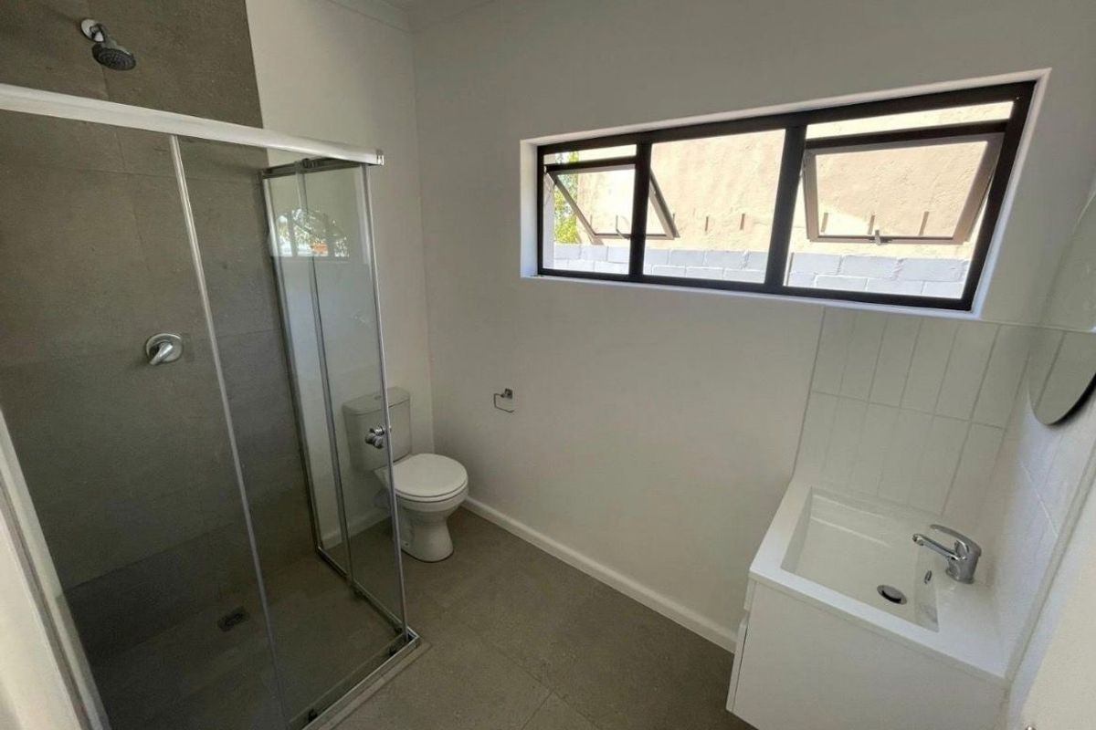 A clean, modern bathroom featuring a glass-enclosed shower, a wall-mounted vanity basin, and neutral floor tiling with a functional top-hung window.