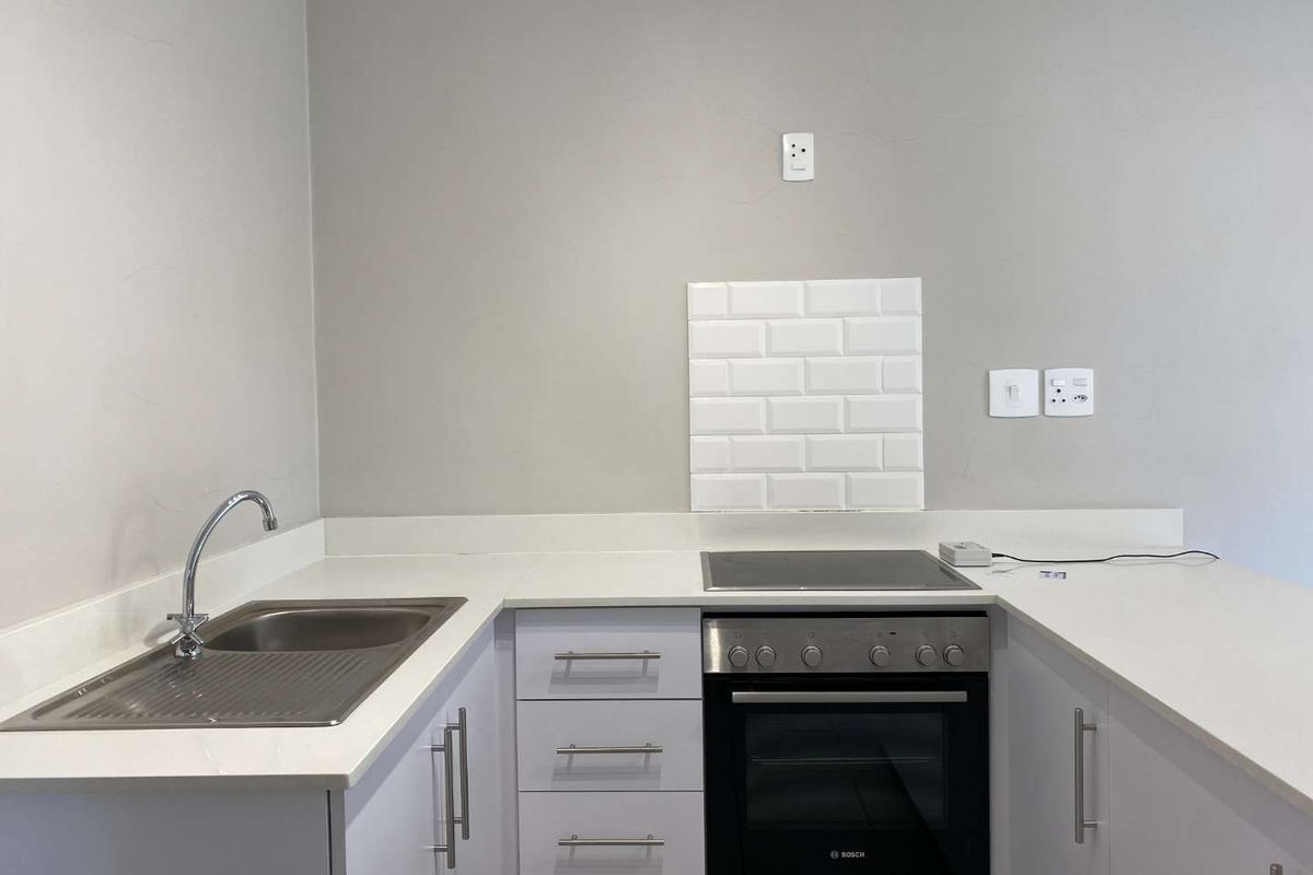 A clean, modern kitchen featuring white cabinetry, stone countertops, a built-in oven, electric hob, and a stainless steel sink with a subway tile splashback.