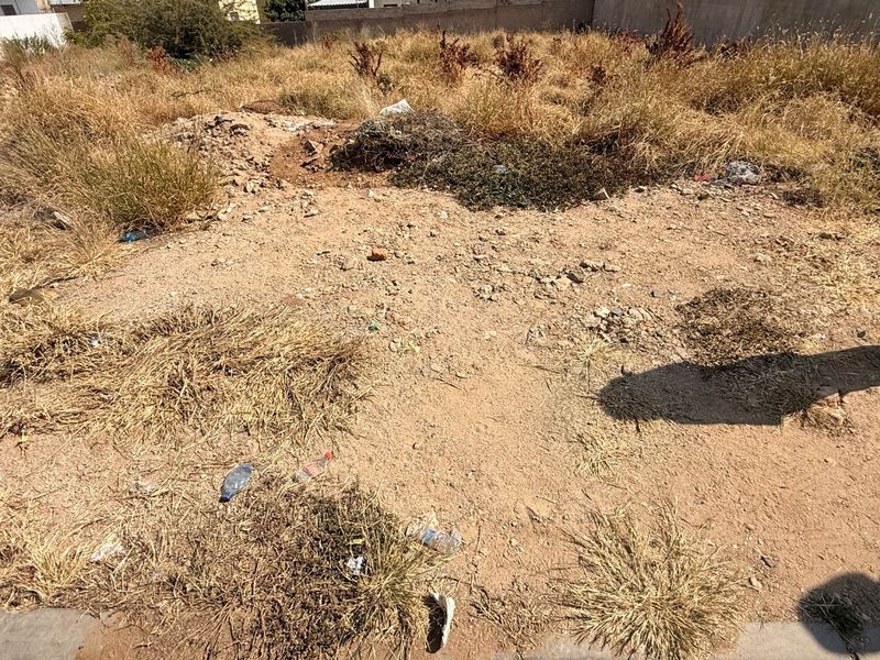 A vacant residential plot featuring dry, overgrown vegetation and a perimeter boundary wall, suitable for future development.