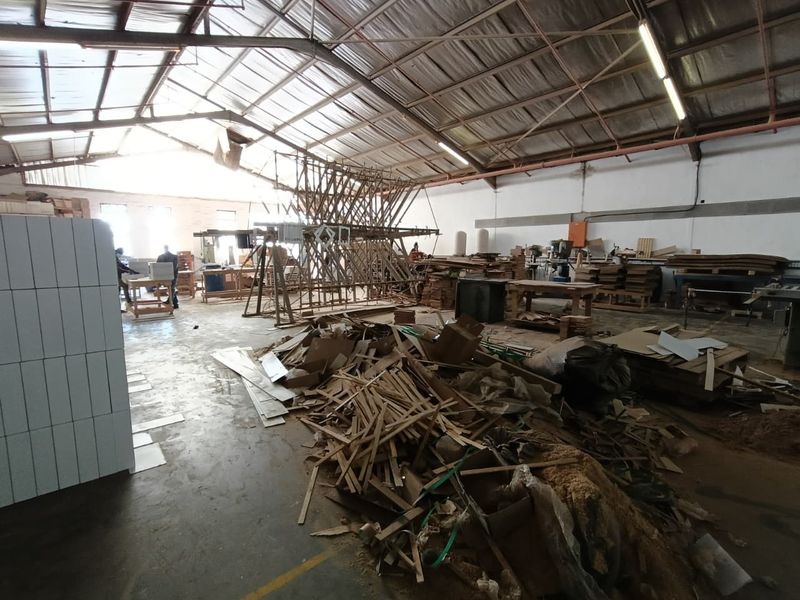 Spacious industrial workshop area featuring exposed steel roof trusses, concrete flooring, and high-bay fluorescent lighting, currently utilized for manufacturing or storage purposes.