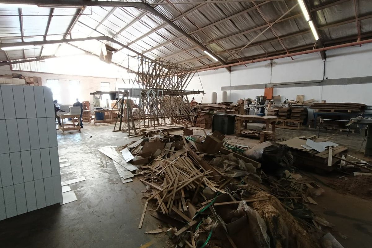 Spacious industrial workshop area featuring exposed steel roof trusses, concrete flooring, and high-bay fluorescent lighting, currently utilized for manufacturing or storage purposes.