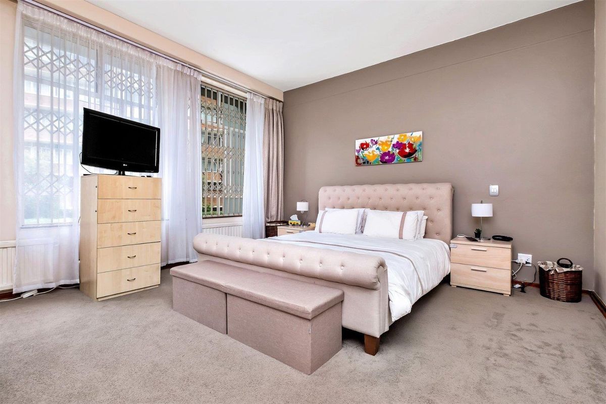 A spacious bedroom featuring neutral wall tones, carpeted flooring, and large windows fitted with security bars and curtains.