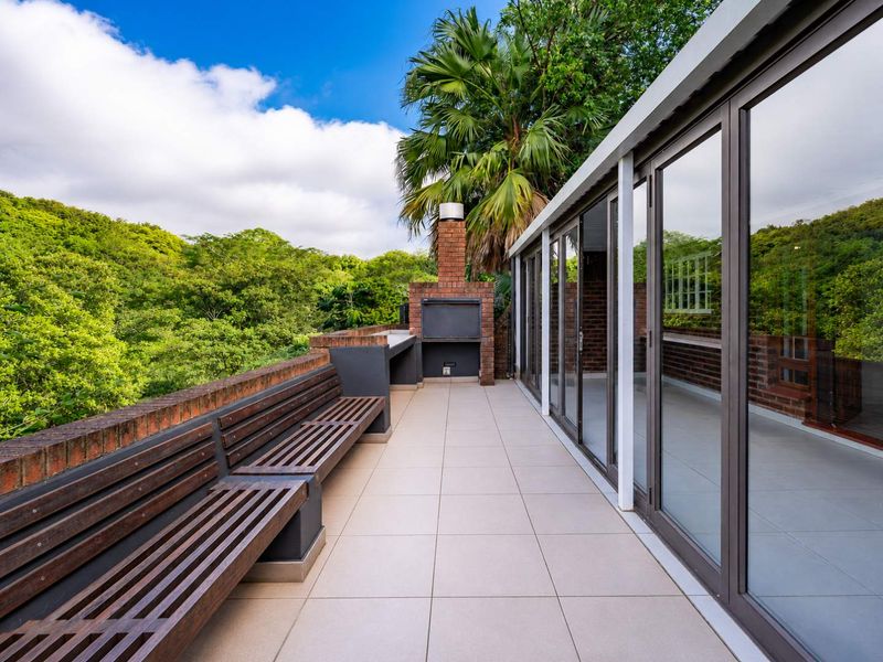 Spacious outdoor balcony featuring a built-in brick braai, integrated wooden seating, and large sliding glass doors overlooking lush greenery.