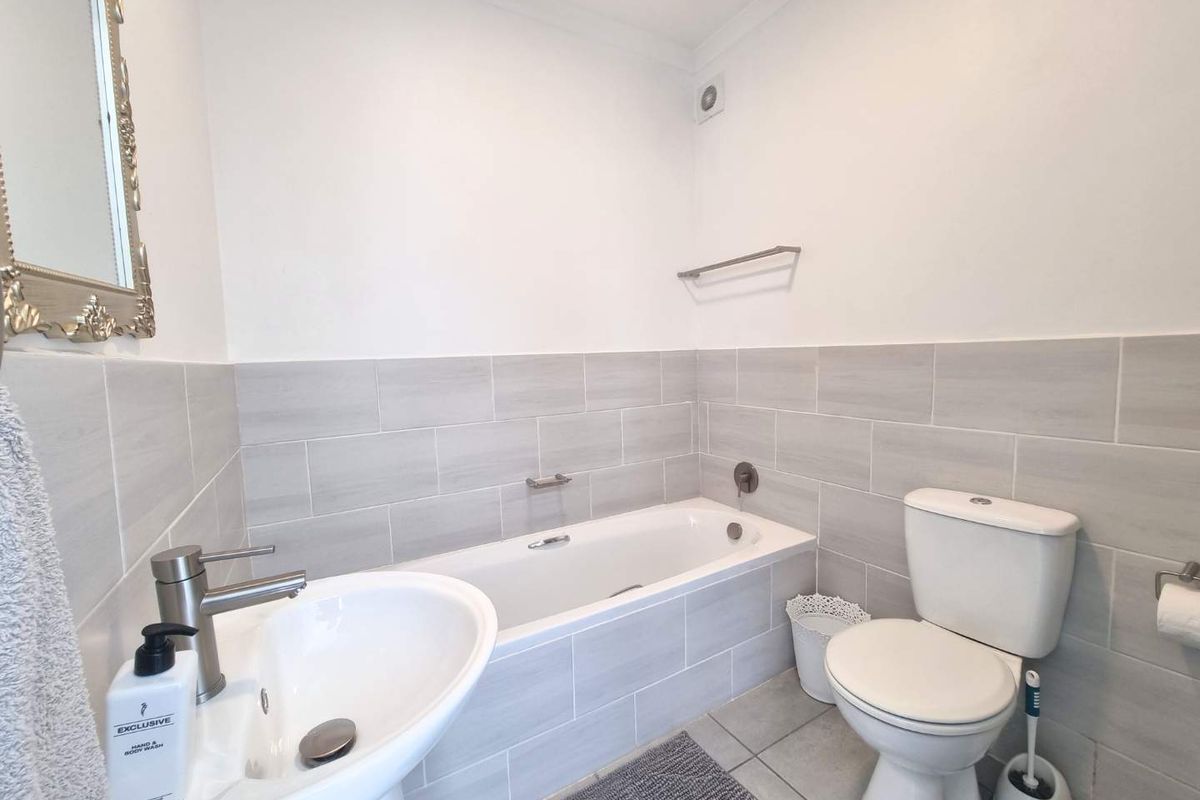A clean, modern bathroom featuring a white bathtub, pedestal sink, and toilet with neutral grey wall tiling and chrome fixtures.