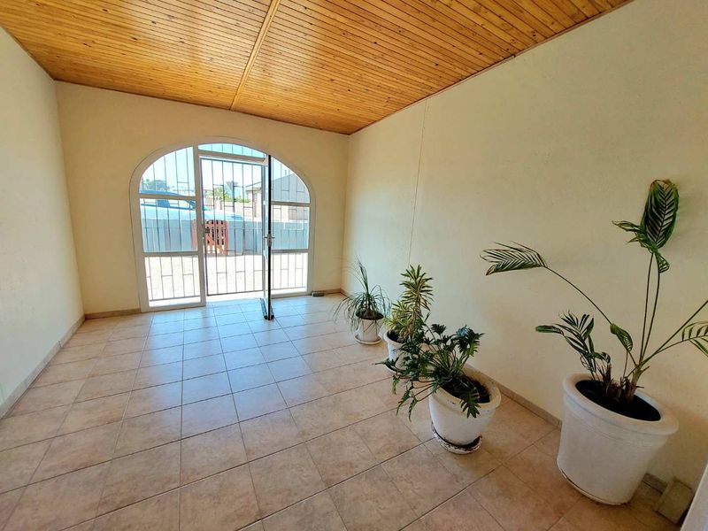 A covered patio area featuring a timber tongue and groove ceiling, ceramic floor tiles, and an arched glass entrance door with a security gate.