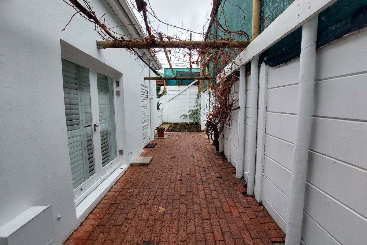 A narrow rear courtyard featuring red brick paving, a timber pergola structure, and white boundary walls with access via french doors.