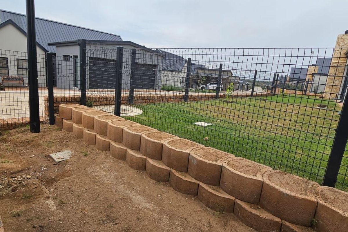 Rear garden area featuring a tiered retaining wall, wire mesh fencing, and a lawn, with adjacent paved driveway and residential building exterior.