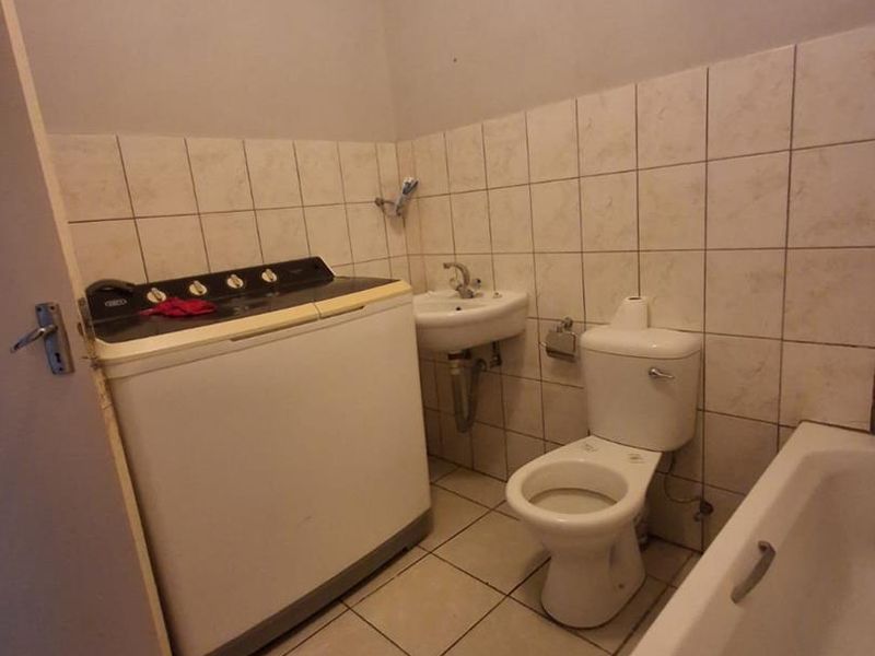 A functional bathroom featuring a bathtub, toilet, and pedestal basin with tiled walls and flooring, currently accommodating a top-loading washing machine.