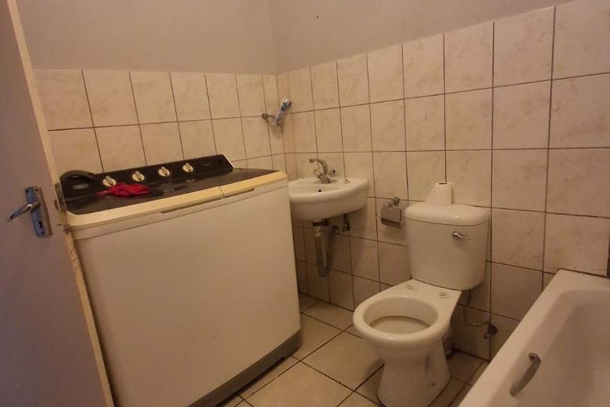 A functional bathroom featuring a bathtub, toilet, and pedestal basin with tiled walls and flooring, currently accommodating a top-loading washing machine.
