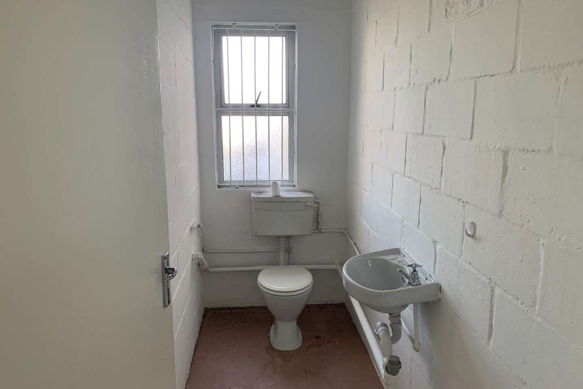A functional industrial-style bathroom featuring a toilet, wall-mounted basin, and security-barred window with exposed brick walls.