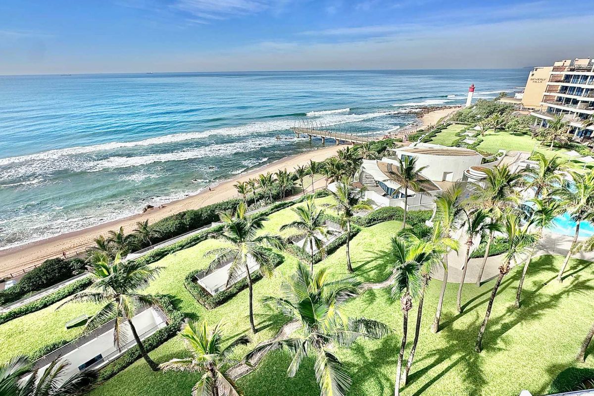 A high-angle aerial view showcasing the coastal landscape, featuring landscaped gardens, a swimming pool area, and direct access to the beach and ocean.