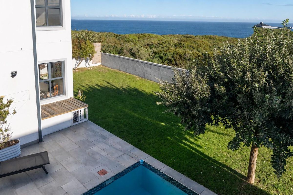 Elevated view of a rear garden featuring a paved patio, swimming pool, and expansive ocean views beyond the boundary wall.