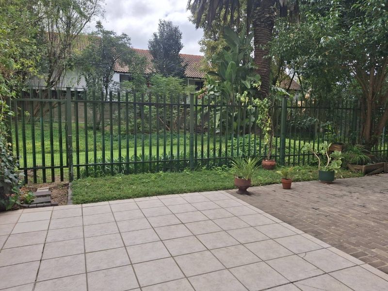 A paved outdoor patio area featuring metal security fencing and established garden surroundings, providing a functional space for outdoor relaxation.